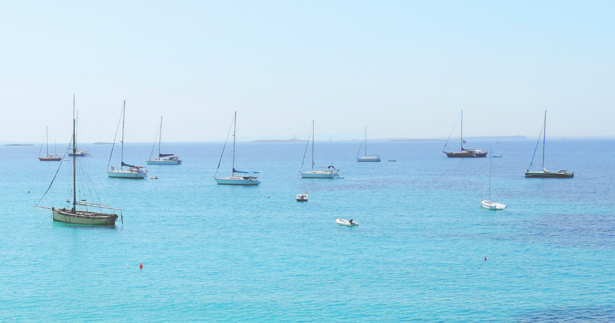 Multiple sailboats and a few smaller boats are anchored in clear turquoise water under a bright sky.